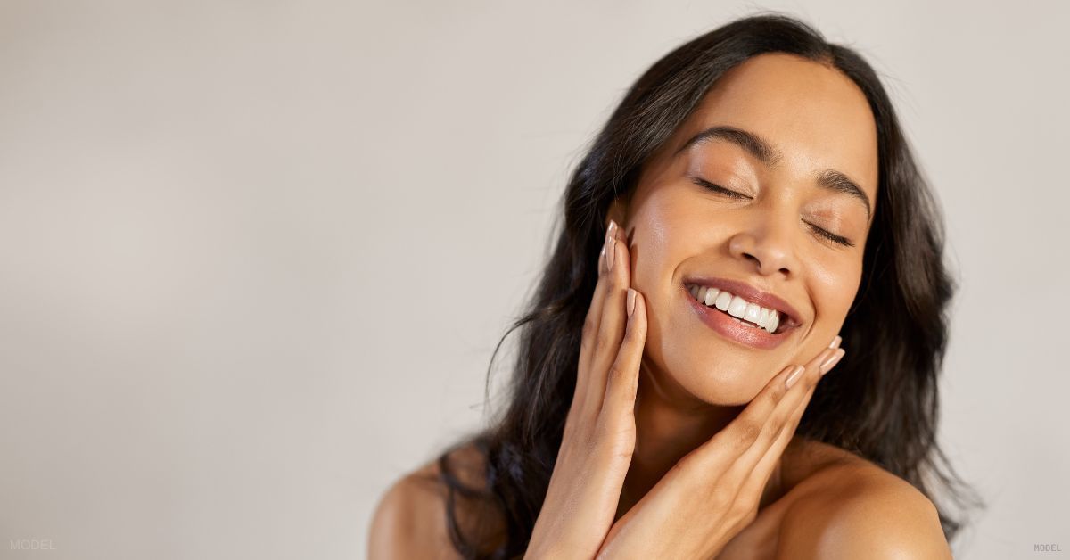 A woman with clear skin, smiling with their hands on their face. (Model)